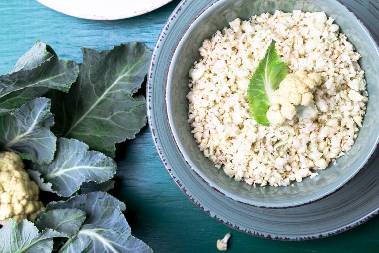 Cauliflower rice in a blue bowl on a blue table next to a head of cauliflower with leaves.